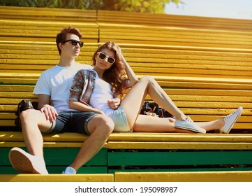 Young Modern Stylish Hipsters Couple Rest On The Bench In City Park, Sunny Portrait Couple Teenagers In Summer Day