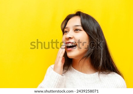 young modest asian girl with braces smiling and taunting on yellow isolated background, korean girl covering her smile and being shy