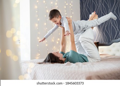 Young Mom In Casual Wear Liying On Bed And Throws Up Happy Little Son While Having Fun Together In Cozy Bedroom At Home. Happy Children With Mother. Christmas Time