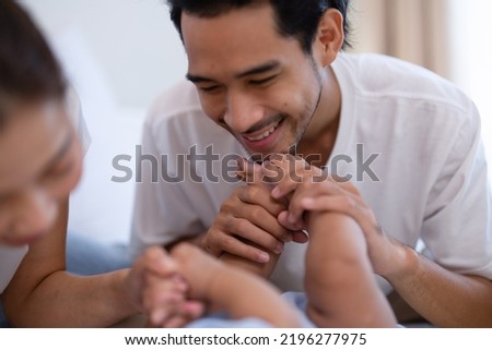 Young Mom and father laughing joyful proud teasing playing kiss cheek and feet arm hold asian infant baby new born on bed, baby have fun looking  to parrent face
