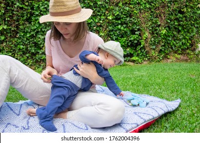 Young Mom In Hat Holding Daughter And Checking Baby Diaper. Little Baby Girl In Blue Clothes Reaching Toy On Plaid. Pretty Mother With Daughter In Park. Summer Family Time And Sunny Days Concept