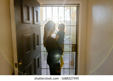 Young Mom Holding Baby Sitting At The Front Entrance With Door Opend Looking Through Metalic Screan Door.