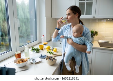 Young Mom Holding Kid While Drinking Water At Home Stock Photo