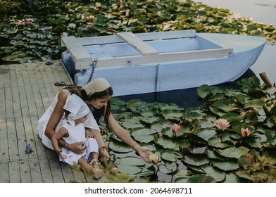 Young mom and little toddler baby girl wearing white light dress are playing on the pier near a lake and a blue boat, watching a lot of flowering waterlilies, family together, water, rural
