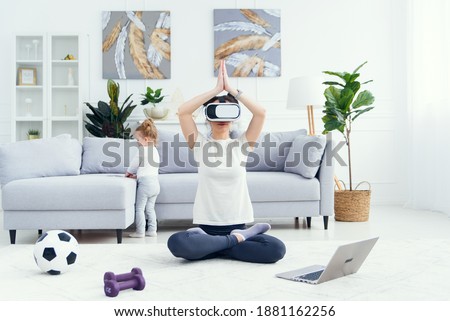 Young mom meditating in lotus yoga position using ar glasses while her daughter watches cartoons on background.