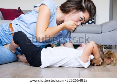 Young mom plays with baby in living room: hides mouth pretending a scary secret and makes big eyes in a funny face that makes her daughter laugh