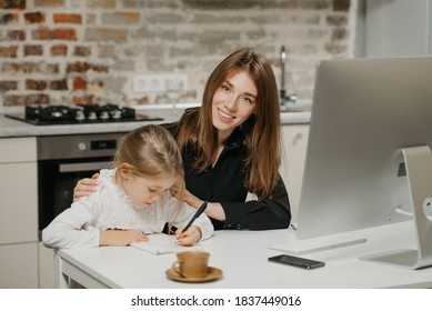 A young mom is posing and hugging a pretty daughter while she is drawing in the notebook at the workplace. A gorgeous mother watches her blonde child which is doing homework at home. - Powered by Shutterstock