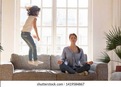 Young mom sit on couch at home meditating in lotus position, small daughter have fun jumping near, concentrated calm mother practice yoga relax on sofa ignore playful kid. Stress free concept - Shutterstock ID 1419584867