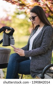 Young Mom Sitting On The Bench With Her Baby In Stroller And Use Phone. Mothe Look Something In Internet