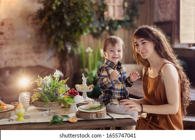 Young Mom And Son Are Sitting At The Festive Table. Easter Treats Decoration Table Setting. Spring Egg Celebration. European Appearance. Country Boho Style Village Toddler Happiness Laugh Family