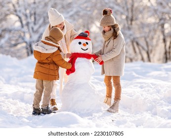 Young mom and two kids making snowman in winter park outdoors. Joyful little brother and sister in warm clothes happily helping mother decorate snow figure with carrot nose and hands with dry branches - Powered by Shutterstock