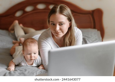 Young mom works remotely on laptop, baby child lies on his tummynext to her mother on bed in bedroom. Concept working mother, remote work, education, maternity leave - Powered by Shutterstock