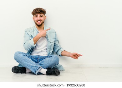 Young Moroccan man sitting on the floor isolated on white background excited holding a copy space on palm.