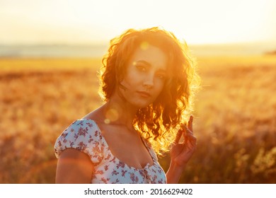 young moroccan woman, with brown curly hair, standing in a wheat field, while the sun ist setting in the background and blinding the camera
