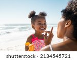 Young mother applying protective sunscreen on daughter nose at beach with copy space. Black woman hand putting sun lotion on female child face. African american cute little girl with sunblock cream.