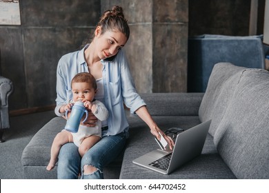 Young mother with baby boy remote working and using laptop during conversation on smartphone