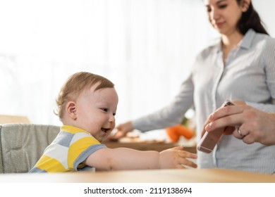 Young Mother And Baby Boy Video Call Online With Family While Eating. Caucasian Beautiful Mom Use Mobile Cell Phone And Chat With Husband While Little Toddler Kid Having Breakfast In Kitchen In House.