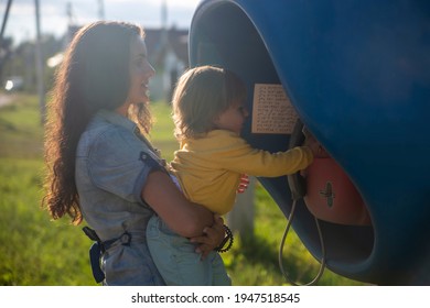 Young Mother And Baby Call On A Landline Street Phone In Telephone Booth In The Village In Summer. Walking With Family Summer Sunny Day In Sunshine. Artistic Focus