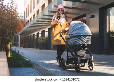 Young mother with baby child laying in stroller walking through fashionable living quarter. Modern urban multistoried building with comfortable apartments. Woman using mobile phone and wifi connection