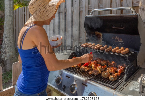 The young mother bastes the chicken\
on the grill with barbecue sauce. She is wearing a broad-rimmed hat\
while holding a bowl of BBQ sauce and a cooking\
brush.