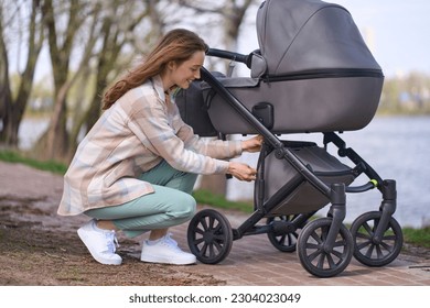 Young mother, beautiful Caucasian red haired woman in casual clothes, opening the trunk in a baby stroller during a walk with her newborn child in the park by river - Powered by Shutterstock