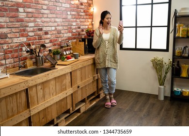 Young Mother Calls Relatives On Video Communication. Full Length Beautiful Elegant Asian House Wife Waving Hand Saying Hi Talking Chatting On Mobile Phone Standing Next To Window In Wooden Kitchen.