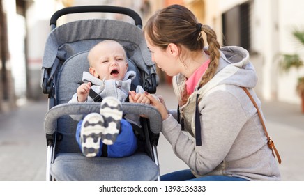 Young Mother Calming A Crying Child Sitting In A Stroller While Walking Down The Street