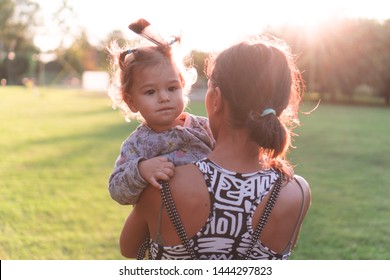 Young Mother Carrying Kid In Arms. Mom Holding Child Outdoors In Urban Park In Golden Hour. Woman Hugging Toddler With Feather In Hair Walking Away From Camera In Nature At Sunset.