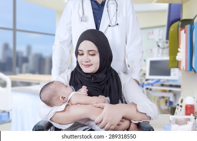 Young Mother Carrying Newborn Baby While Sitting On Wheelchair With Her Doctor Pushing It