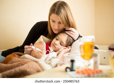 Young Mother Checking Temperature Of Sick Daughter Lying In Bed