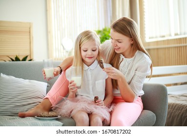 Young Mother With Child Daughter At Home On The Couch Drinking Milk And Cookies, Laughing And Talking Together