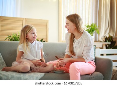 Young Mother With Child Daughter At Home On The Couch Drinking Milk And Cookies, Laughing And Talking Together