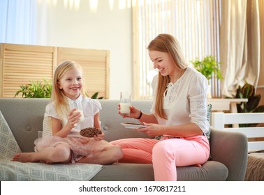 Young Mother With Child Daughter At Home On The Couch Drinking Milk And Cookies, Laughing And Talking Together