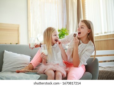 Young Mother With Child Daughter At Home On The Couch Drinking Milk And Cookies, Laughing And Talking Together