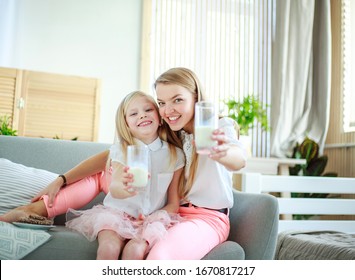 Young Mother With Child Daughter At Home On The Couch Drinking Milk And Cookies, Laughing And Talking Together