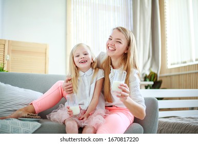 Young Mother With Child Daughter At Home On The Couch Drinking Milk And Cookies, Laughing And Talking Together