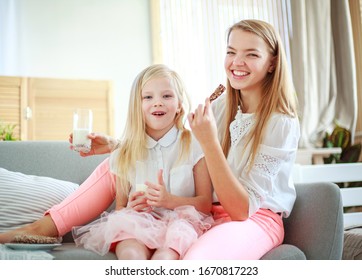 Young Mother With Child Daughter At Home On The Couch Drinking Milk And Cookies, Laughing And Talking Together