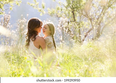 Young Mother And Child Daughter Together, Hugging And Joyfully Laughing While Relaxing In A Golden Field Of Sunshine And Spring Flowers While On A Summer Holiday. Family Outdoors Lifestyle.