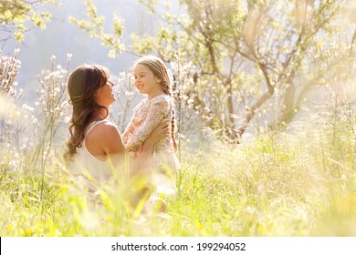 Young Mother And Child Daughter Together, Hugging And Smiling Sitting And Relaxing In A Golden Field Of Sunshine And Spring Flowers While On A Summer Holiday. Family Activities And Outdoors Lifestyle.