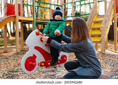 Young mother with child playing on playground, mom holding hand of laughing cheerful baby kid swinging on toy horse outdoors. Childhood, motherhood, active recreation for children. - Powered by Shutterstock