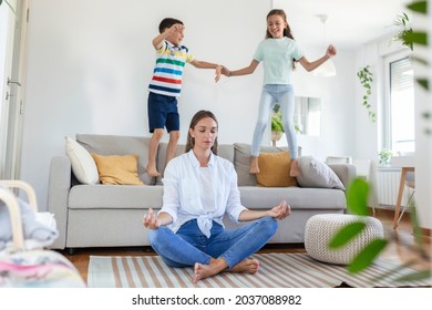 Young mother with closed eyes meditating in lotus pose on floor trying to save inner harmony while excited children jumping on sofa and screaming in light spacious living room - Powered by Shutterstock