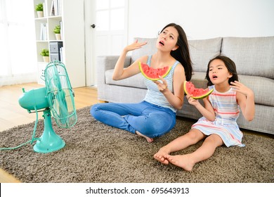 Young Mother With Cute Little Daughter Feeling Hot In Summer Sitting On Living Room Floor Blowing Electric Fan Refreshing And Eating Cold Watermelon.