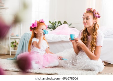 young mother and cute little daughter with curlers in tutu tulle skirts having tea party together - Powered by Shutterstock