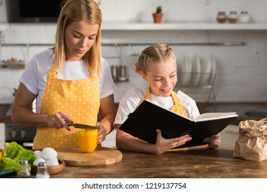 Young Mother Cutting Pepper And Smiling Daughter Reading Cookbook 