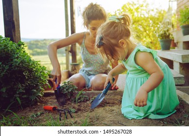 Young Mother And Daughter Digging Picking Ground Dirt Land In Their Yard Garden Near The Home House Family Planting Flowers Or Seeds Using Gardening Tools In Summer Day