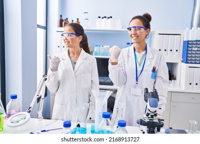 Young Mother And Daughter At Scientist Laboratory Smiling With Happy Face Looking And Pointing To The Side With Thumb Up. 