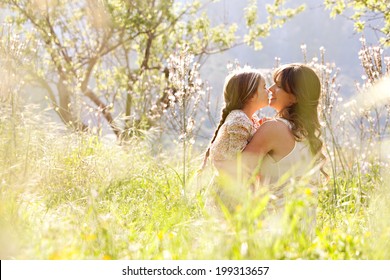 Young Mother And Daughter Together, Hugging And Kissing While Sitting And Relaxing In A Golden Field Of Sunshine And Spring Flowers While On A Summer Holiday. Family Activities And Outdoors Lifestyle.