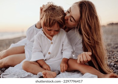 A young mother and daughter in white dresses are sitting on a rocky ocean shore at sunset. A little daughter looks at a stone in her hands, and her mother kisses her - Powered by Shutterstock