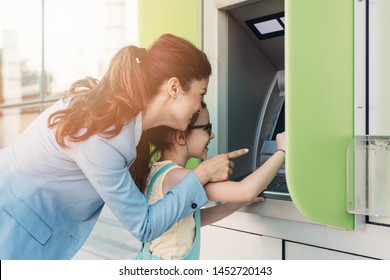 Young mother elegantly dressed with her daughter using ATM machine on city street.