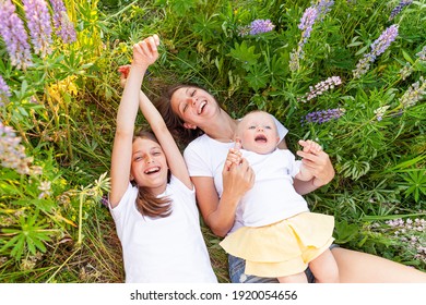 Young Mother Embracing Her Kids Outdoor. Woman Baby Child And Teenage Girl Lying Down On Summer Field With Blooming Wild Flowers Green Background. Happy Family Mom And Daughters Playing On Meadow